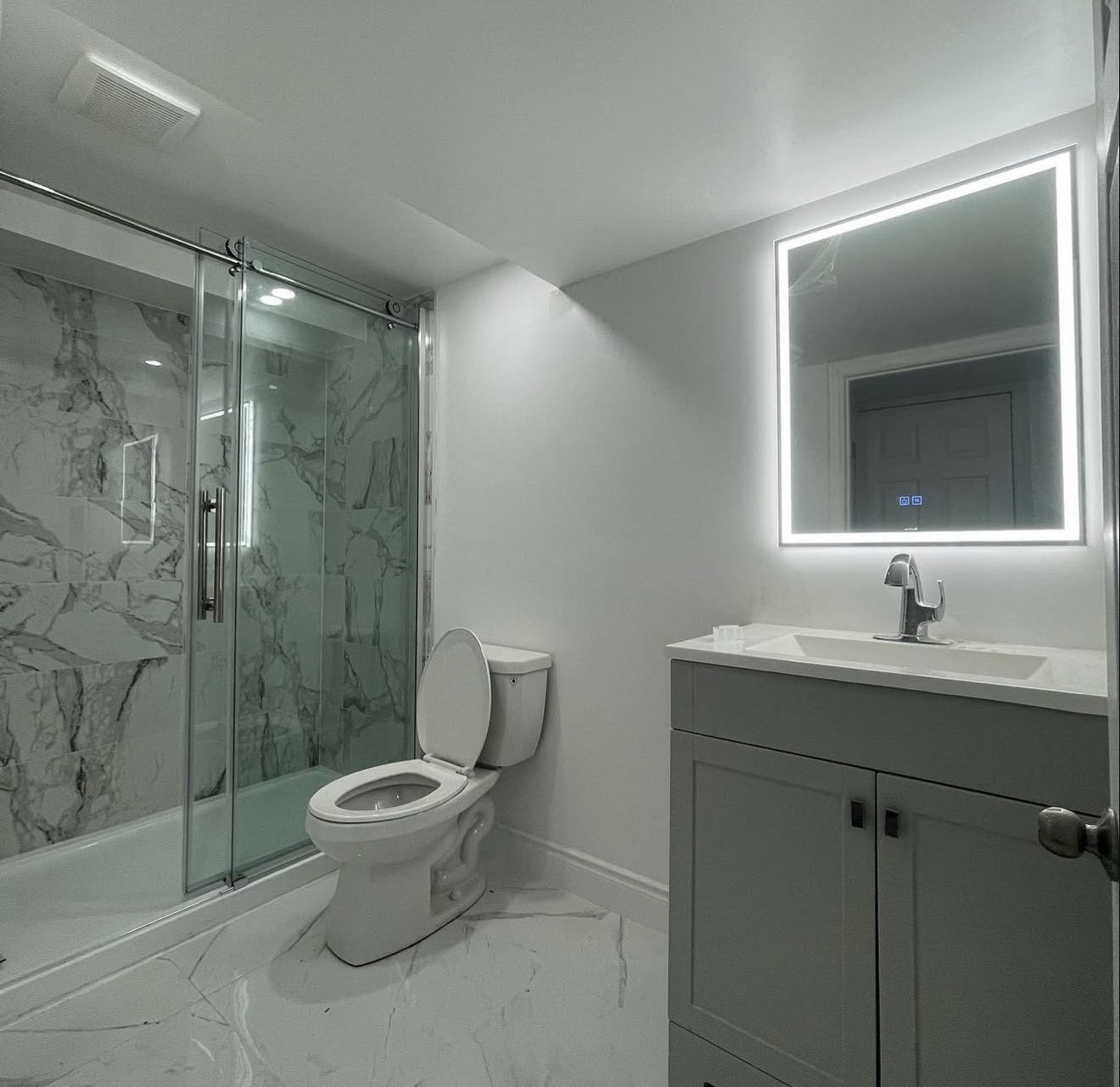 Lower level bathroom with marble-look tile, glass shower and backlit mirror
